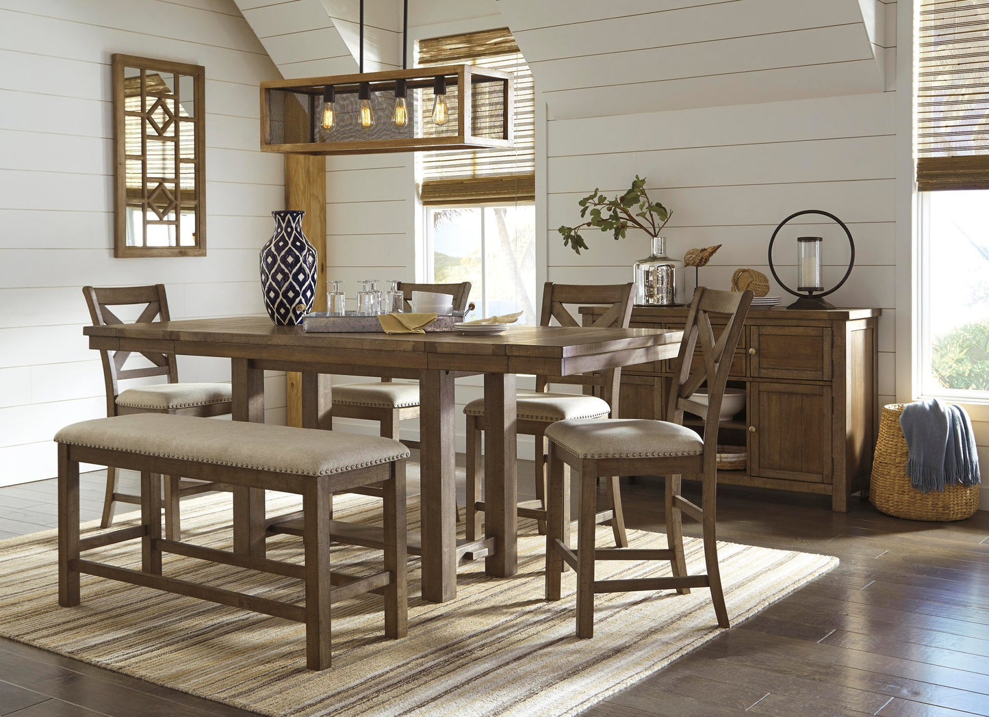 A rustic dining room showcases a wooden table with the Moriville Double UPH Bench, matching chairs, a patterned rug, sideboard, window blinds, and a geometric pendant light—all accentuated by the table’s distressed nutmeg finish.