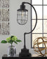 The Jae Metal Desk Lamp (1/CN), in black with a curved arm, sits on a table beside a glass vase of green flowers and a decorative metal sphere, with a window in the background.