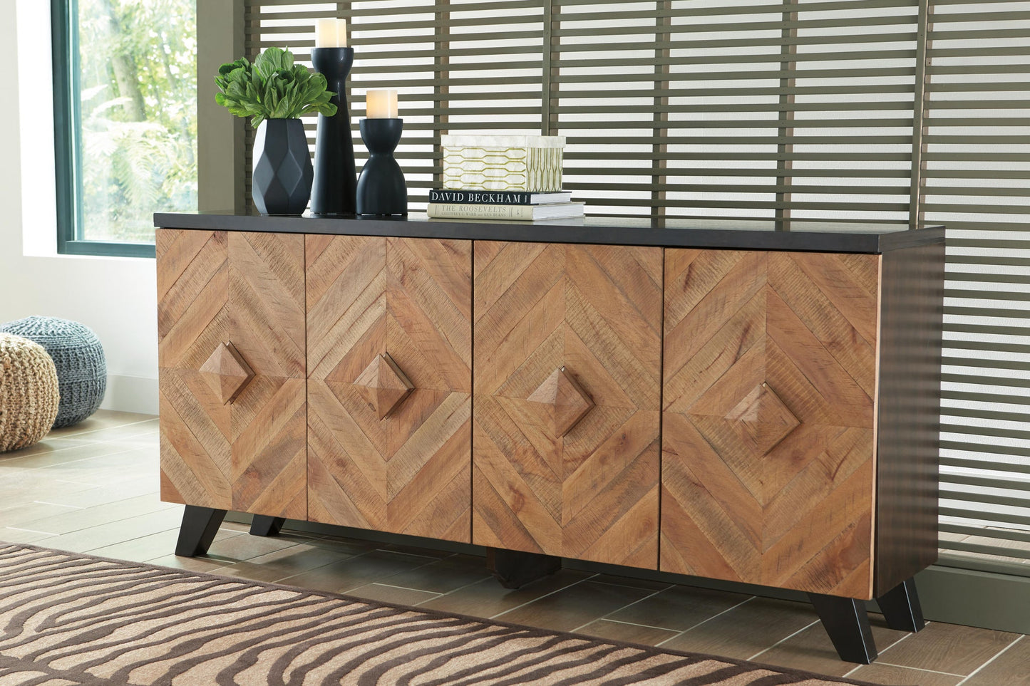 The Robin Ridge Accent Cabinet, featuring two-tone wood and geometric patterned doors with a black top, is styled with candles, vases, and stacked books against a window and slatted wall.