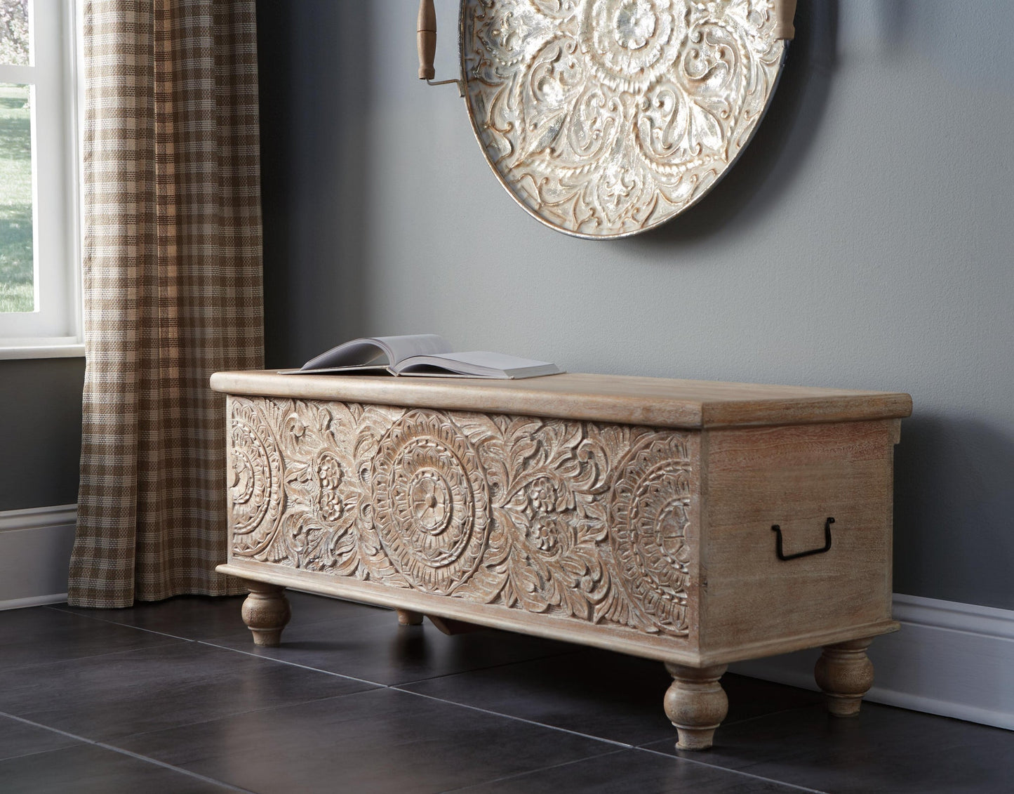 The Fossil Ridge Storage Bench, featuring an antique white finish and ornate hand-carved details, sits on dark tile flooring beneath a round wall piece near a window with plaid curtains. An open book rests on top.