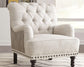 The Tartonelle Accent Chair, featuring button tufting, rolled arms, nailhead trim, and dark wooden legs, sits on a patterned rug in a bright room.