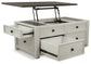 The Bolanburg Lift Top Cocktail Table features an antique white finish, a dark wood top with metal hinges that lifts to reveal extra compartments, and multiple open storage drawers.