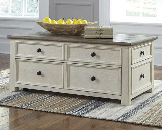 The Bolanburg Lift Top Cocktail Table with storage drawers, a tray of yellow pears, and three books sits on a striped rug in a bright room, featuring an elegant antique white finish.