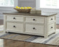 The Bolanburg Lift Top Cocktail Table with storage drawers, a tray of yellow pears, and three books sits on a striped rug in a bright room, featuring an elegant antique white finish.