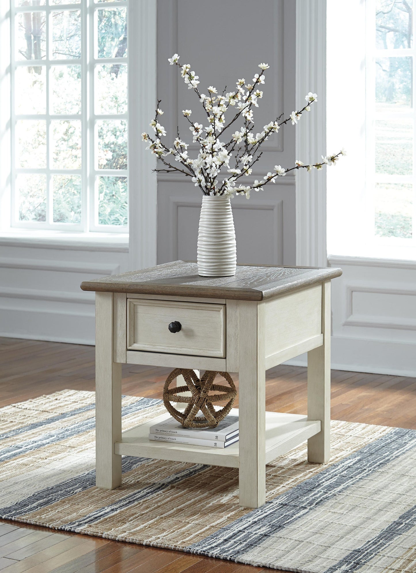 The Bolanburg Rectangular End Table, with its two-tone distressed finish, features a smooth-gliding drawer and shelf. It sits on a striped rug and is topped with a white vase of flowers in a bright, sunlit room.