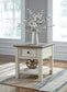 The Bolanburg Rectangular End Table, with its two-tone distressed finish, features a smooth-gliding drawer and shelf. It sits on a striped rug and is topped with a white vase of flowers in a bright, sunlit room.