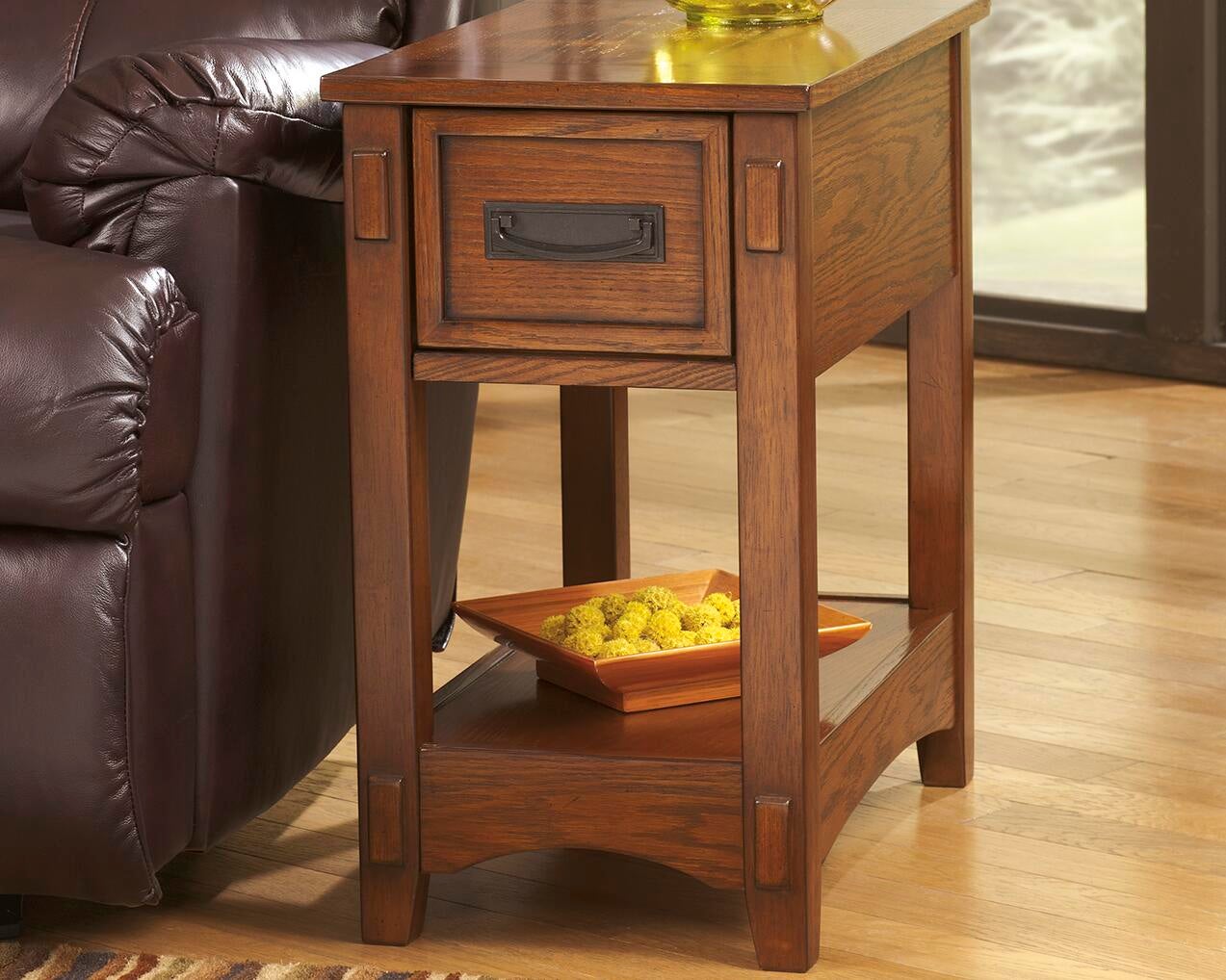 The Breegin Chair Side End Table, featuring a drawer and lower shelf, holds an orange decorative dish with yellow items and stands next to a brown leather sofa on a hardwood floor.