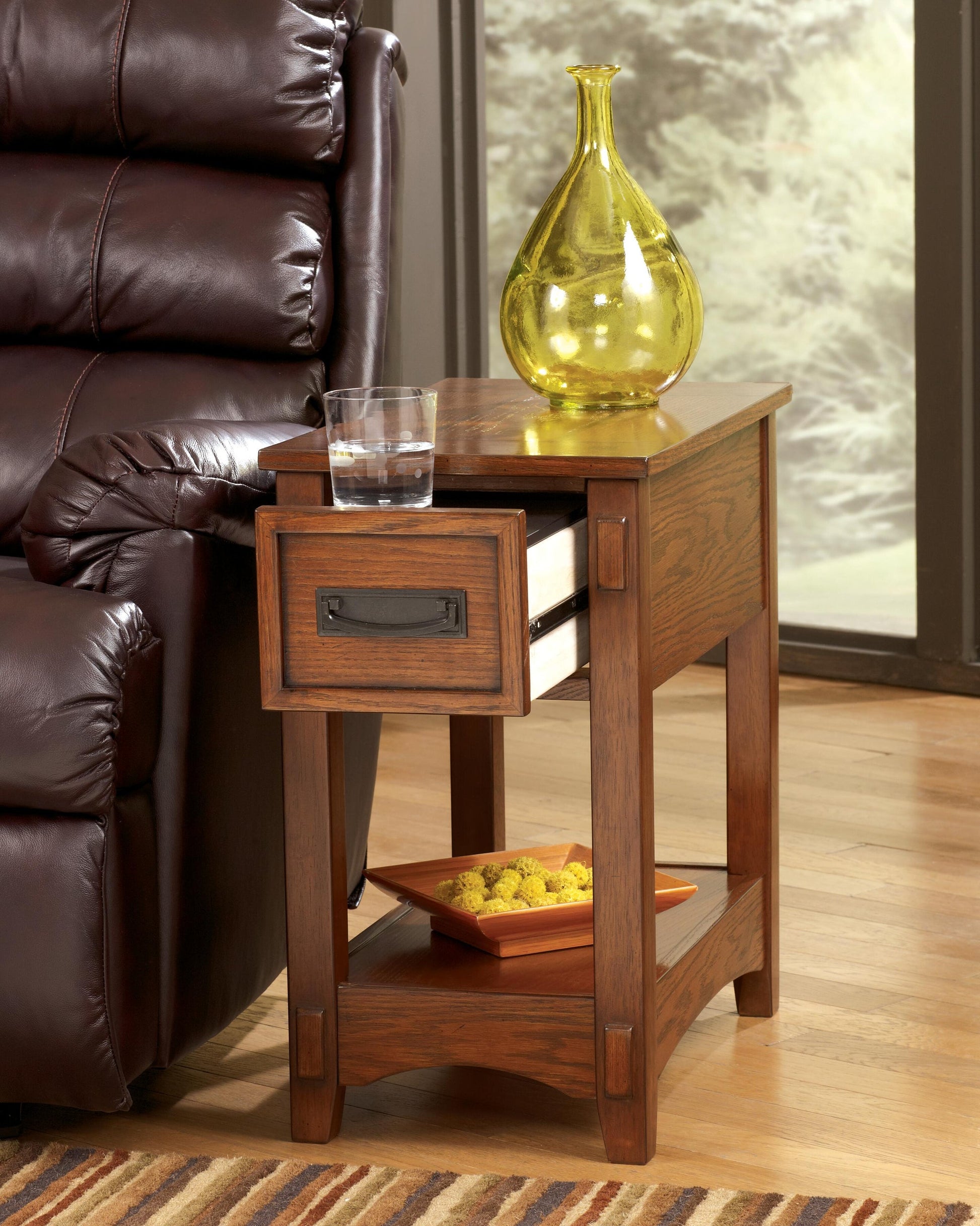 The Breegin Chair Side End Table, featuring an open drawer and lower shelf with a square dish of decorative balls, holds a glass of water and yellow glass vase on top, and stands beside a brown leather chair.