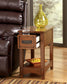 The Breegin Chair Side End Table, featuring an open drawer and lower shelf with a square dish of decorative balls, holds a glass of water and yellow glass vase on top, and stands beside a brown leather chair.