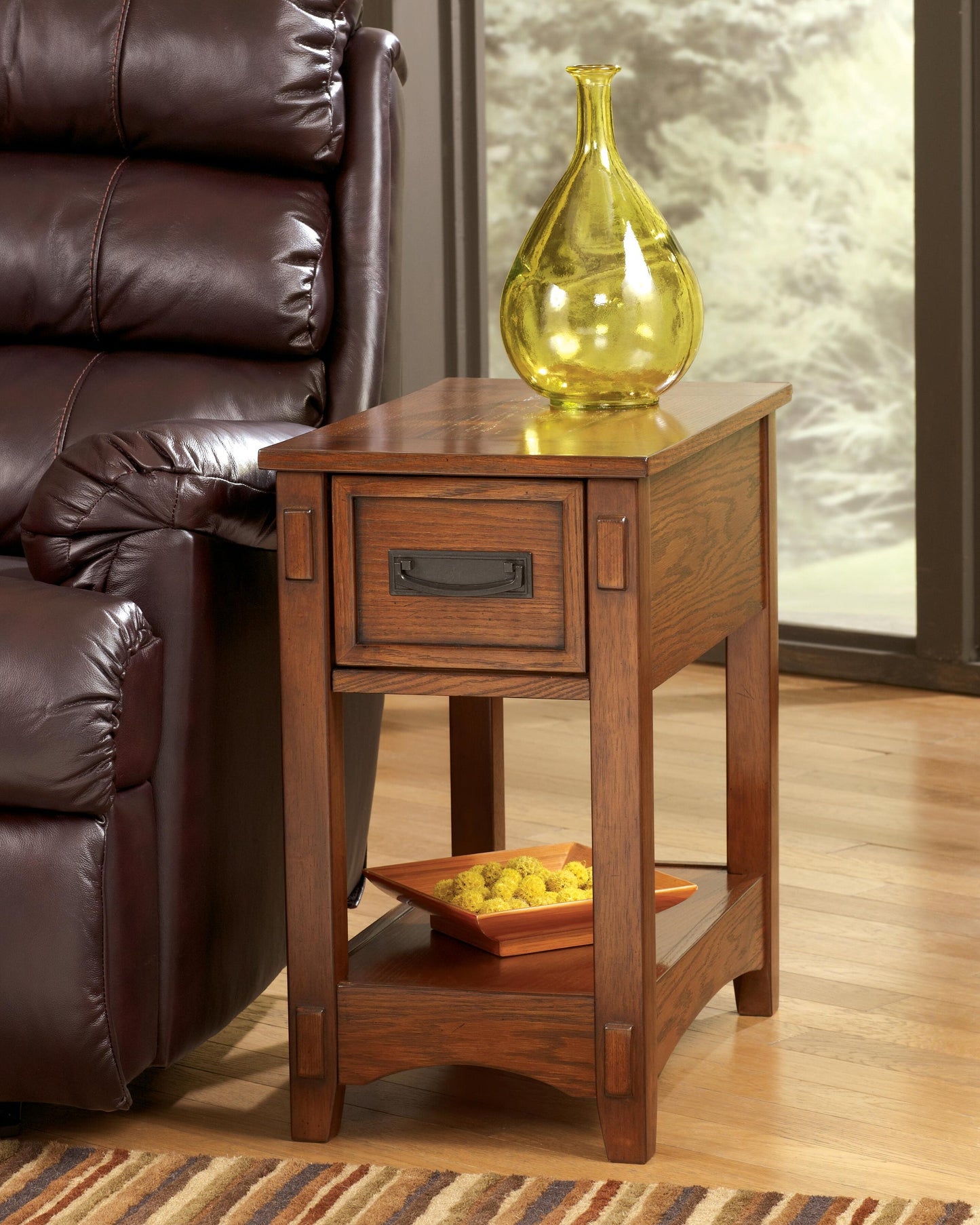The Breegin Chair Side End Table, featuring a drawer and lower shelf, holds a yellow glass vase on top and an orange dish with decorative balls below. It's the perfect accent next to a brown leather sofa in any living space.