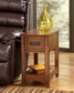 The Breegin Chair Side End Table, featuring a drawer and lower shelf, holds a yellow glass vase on top and an orange dish with decorative balls below. It's the perfect accent next to a brown leather sofa in any living space.