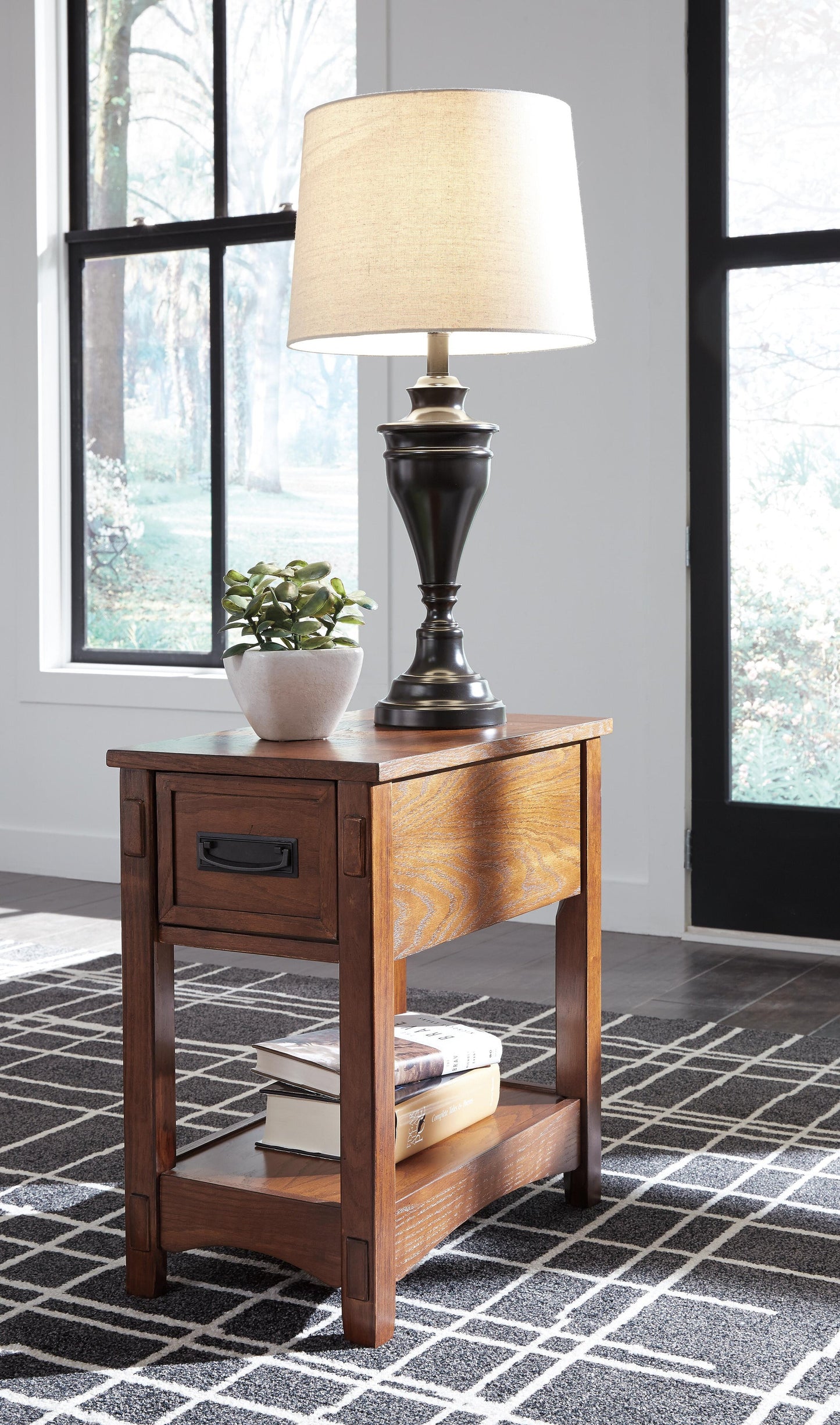 The Breegin Chair Side End Table, featuring a drawer, displays a black lamp, a small potted plant, and stacked books on its lower shelf. This stylish piece rests on a geometric-patterned rug by the window.