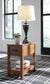 The Breegin Chair Side End Table, featuring a drawer, displays a black lamp, a small potted plant, and stacked books on its lower shelf. This stylish piece rests on a geometric-patterned rug by the window.