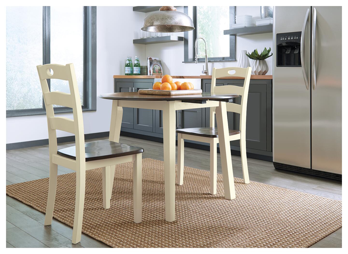 The Woodanville Round DRM Drop Leaf Table with two chairs sits on a woven rug in a modern kitchen, accented by a bowl of oranges and stainless steel appliances.