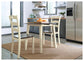 The Woodanville Round DRM Drop Leaf Table with two chairs sits on a woven rug in a modern kitchen, accented by a bowl of oranges and stainless steel appliances.