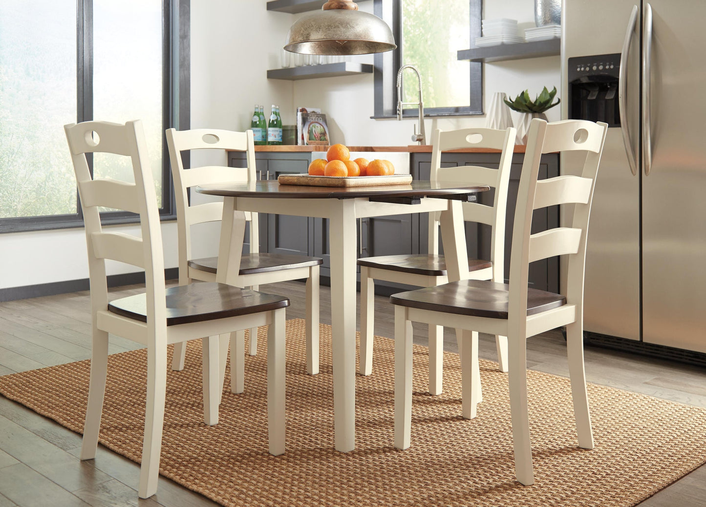 A kitchen featuring the Woodanville Round DRM Drop Leaf Table, four white chairs with dark seats, a bowl of oranges, modern appliances, and a woven rug creates a cozy and inviting space.
