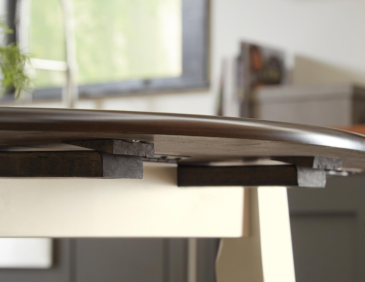 A close-up of a table extension leaf partially extended under the Woodanville Round DRM Drop Leaf Table, with a blurred background.
