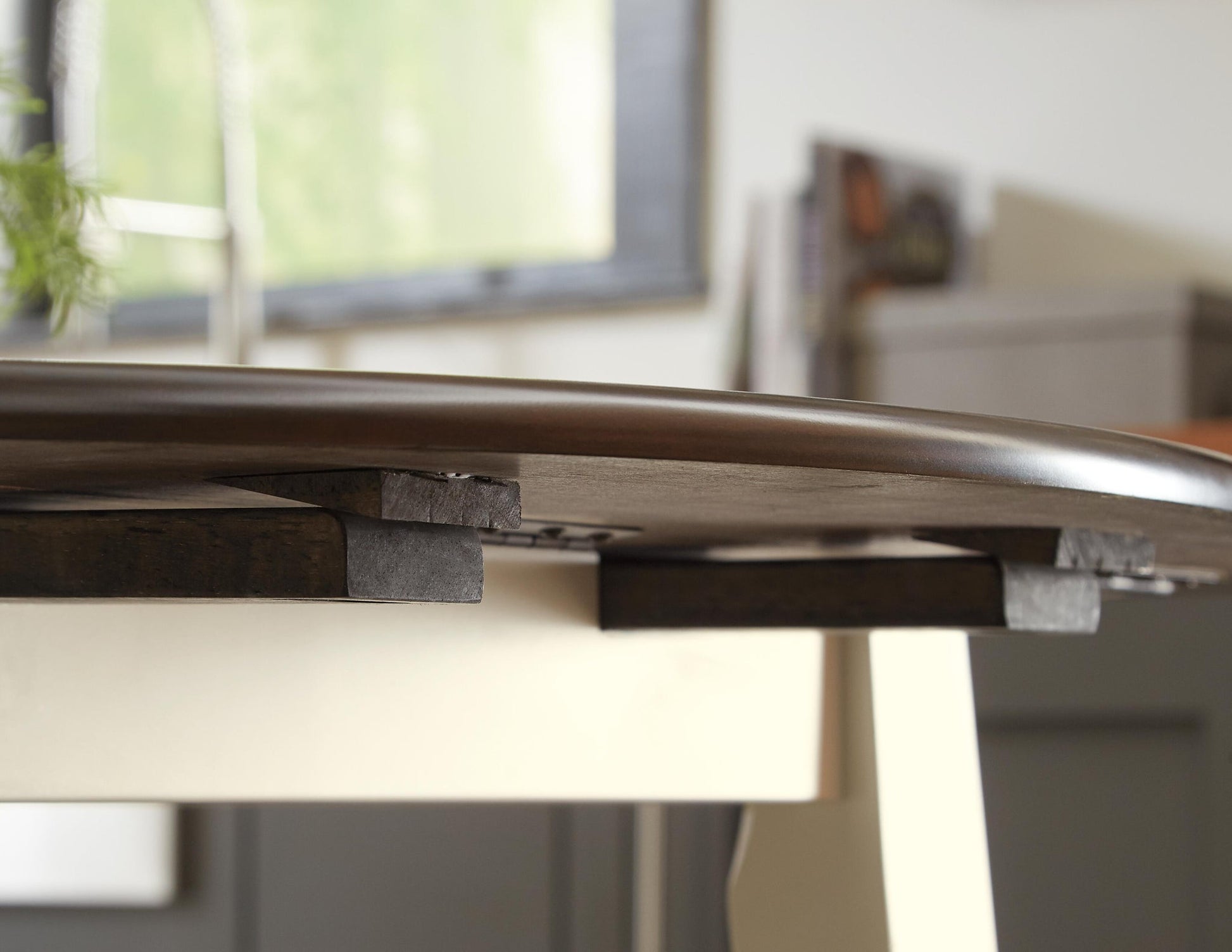 A close-up of a table extension leaf partially extended under the Woodanville Round DRM Drop Leaf Table, with a blurred background.