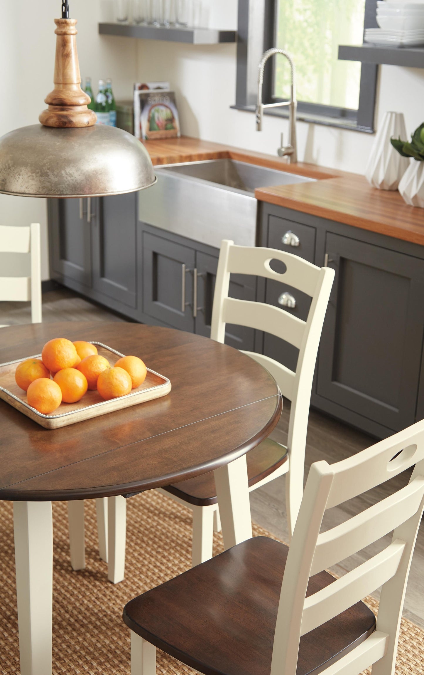 The Woodanville Round DRM Drop Leaf Table, topped with a basket of oranges and surrounded by chairs, sits beside a farmhouse sink and dark gray cabinets beneath a window.