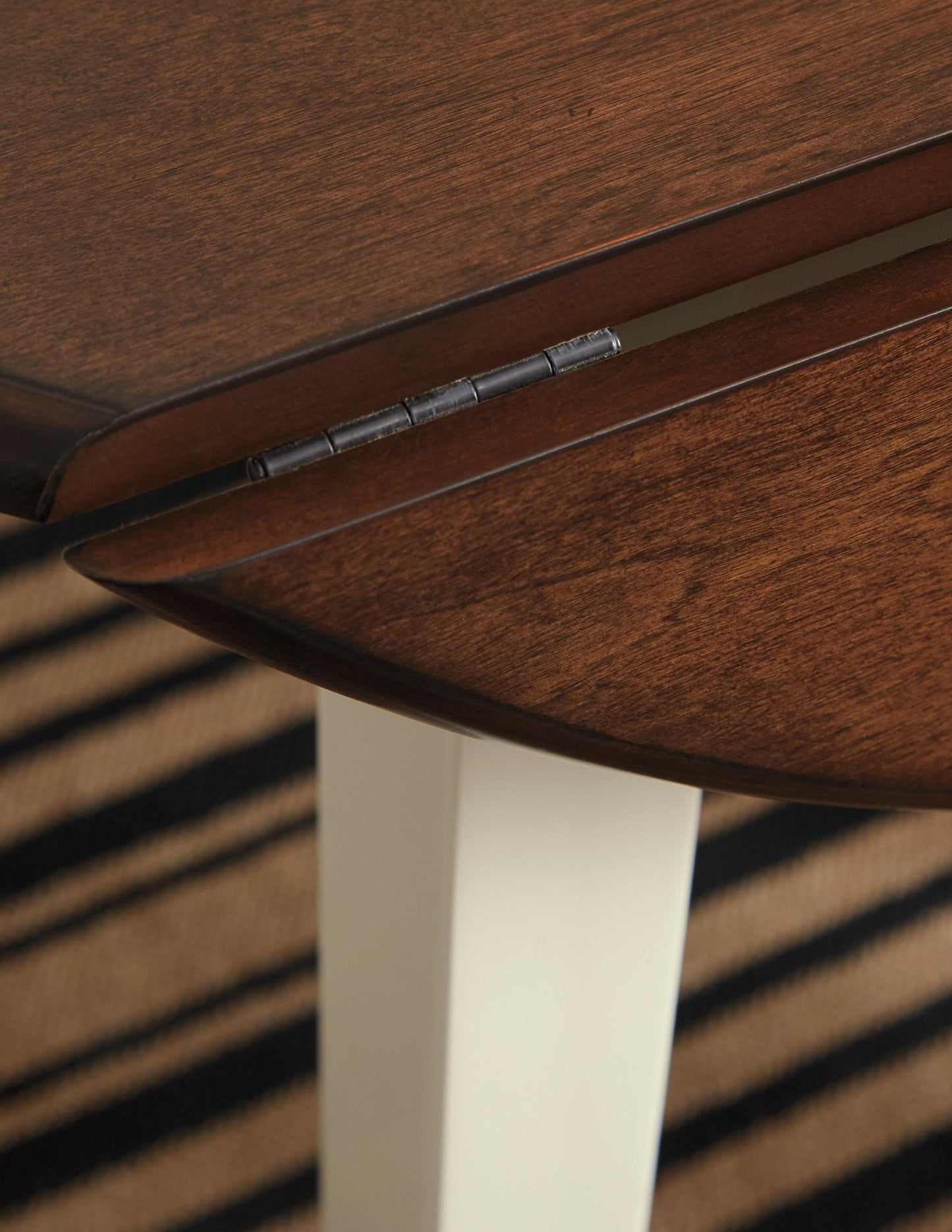 Close-up of the Woodanville Round DRM Drop Leaf Table hinge, featuring a dark brown finish, cream-colored legs, and a striped rug underneath.