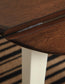 Close-up of the Woodanville Round DRM Drop Leaf Table hinge, featuring a dark brown finish, cream-colored legs, and a striped rug underneath.
