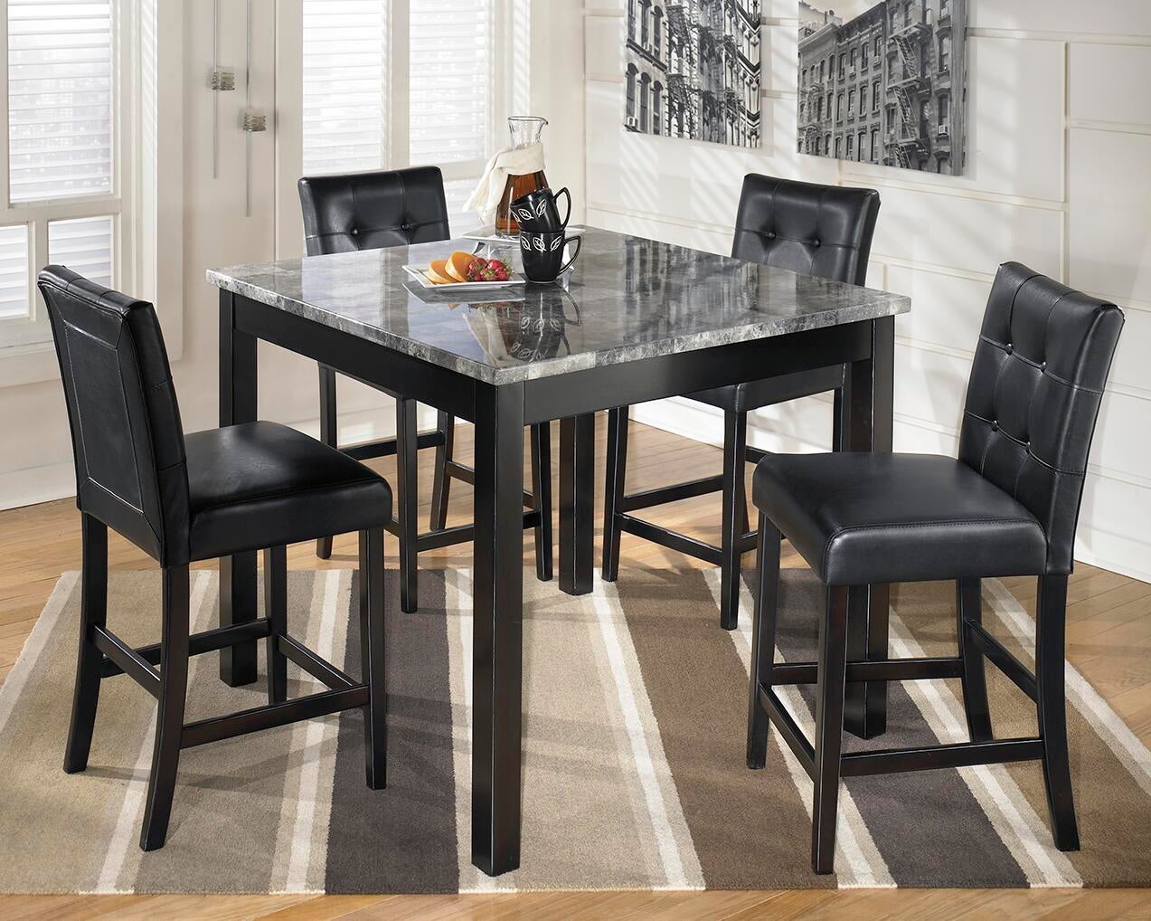 The Maysville Square Counter TBL Set (5/CN), featuring a faux marble tabletop and four black cushioned chairs, sits on a striped rug in a bright room with wall art and large windows.