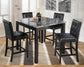 The Maysville Square Counter TBL Set (5/CN), featuring a faux marble tabletop and four black cushioned chairs, sits on a striped rug in a bright room with wall art and large windows.