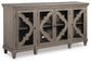 The Fossil Ridge Accent Cabinet is a gray wooden 4-door cabinet with glass inlays, decorative geometric patterns, two adjustable shelves inside, and short legs.