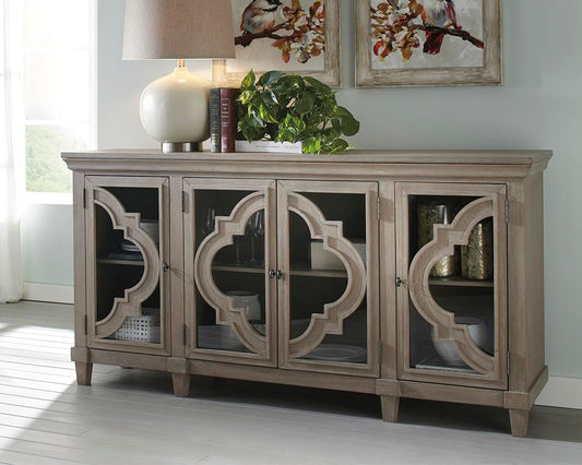 The Fossil Ridge Accent Cabinet is a 4-door sideboard with glass inlays, decorative geometric trim, and adjustable shelves, topped with a lamp and plant, with artwork on the wall behind.