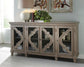 The Fossil Ridge Accent Cabinet is a 4-door sideboard with glass inlays, decorative geometric trim, and adjustable shelves, topped with a lamp and plant, with artwork on the wall behind.