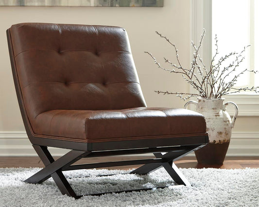 The Sidewinder Accent Chair, crafted in brown faux leather with black metal legs, sits on a white rug beside a ceramic vase of branches in a bright room with light walls and large windows, inspired by the iconic Barcelona chair style.