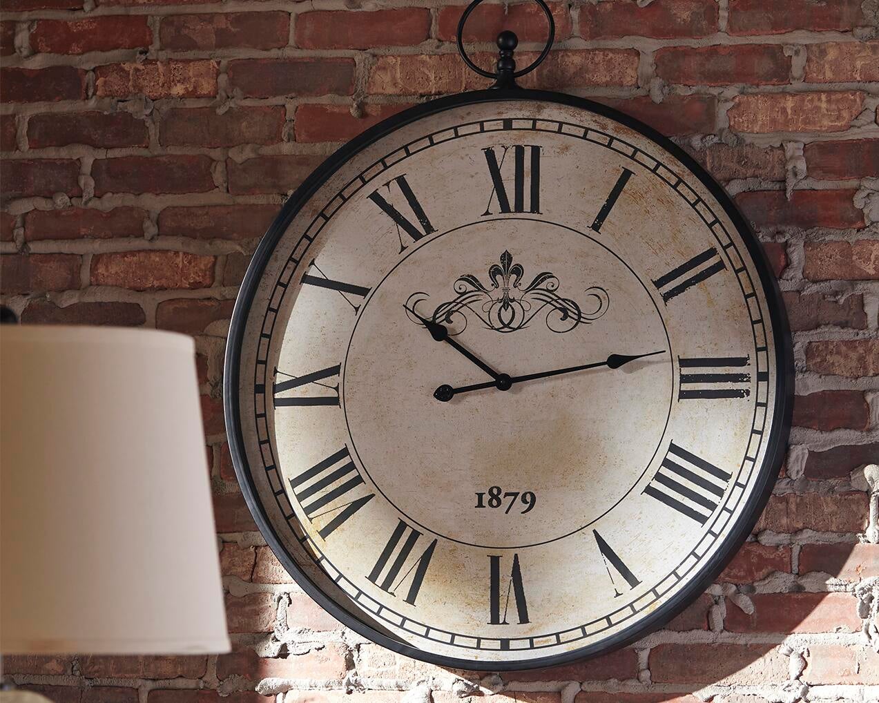 The Augustina Wall Clock, featuring a distressed cream finish and bold Roman numerals reading 2:10, is mounted on a brick wall with part of a lamp in the foreground.