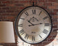 The Augustina Wall Clock, featuring a distressed cream finish and bold Roman numerals reading 2:10, is mounted on a brick wall with part of a lamp in the foreground.