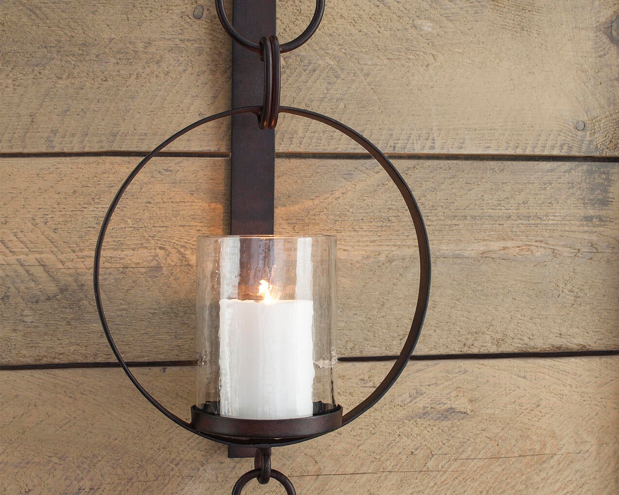 The Ogaleesha Wall Sconce features a white pillar candle in a glass holder, set within a circular bronze-tone metal frame and mounted on wood for an elegant wall accent.
