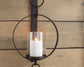 The Ogaleesha Wall Sconce features a white pillar candle in a glass holder, set within a circular bronze-tone metal frame and mounted on wood for an elegant wall accent.