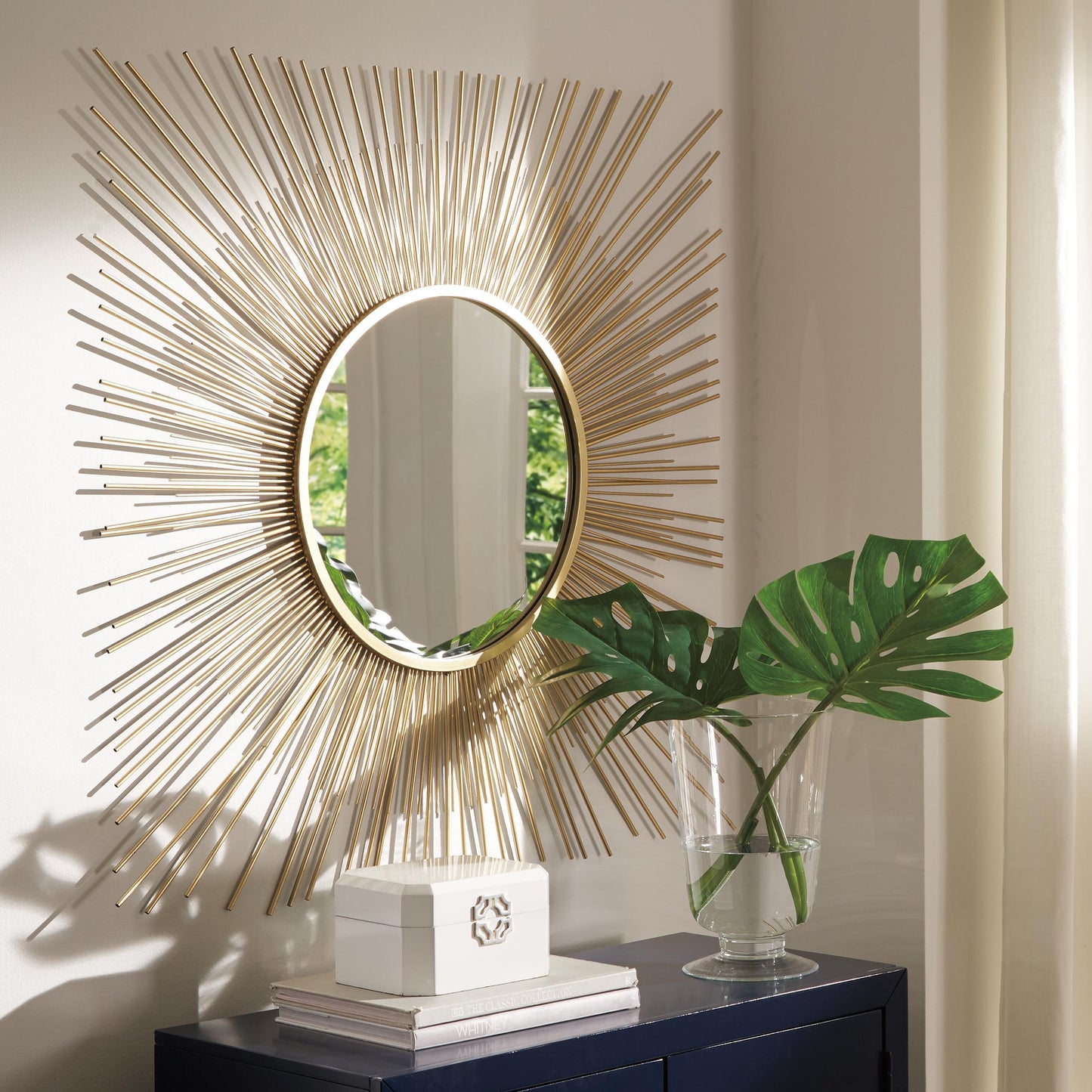 The Elspeth Accent Mirror with a gold sunburst design hangs above a dark cabinet topped with a glass vase of green leaves and a white decorative box.
