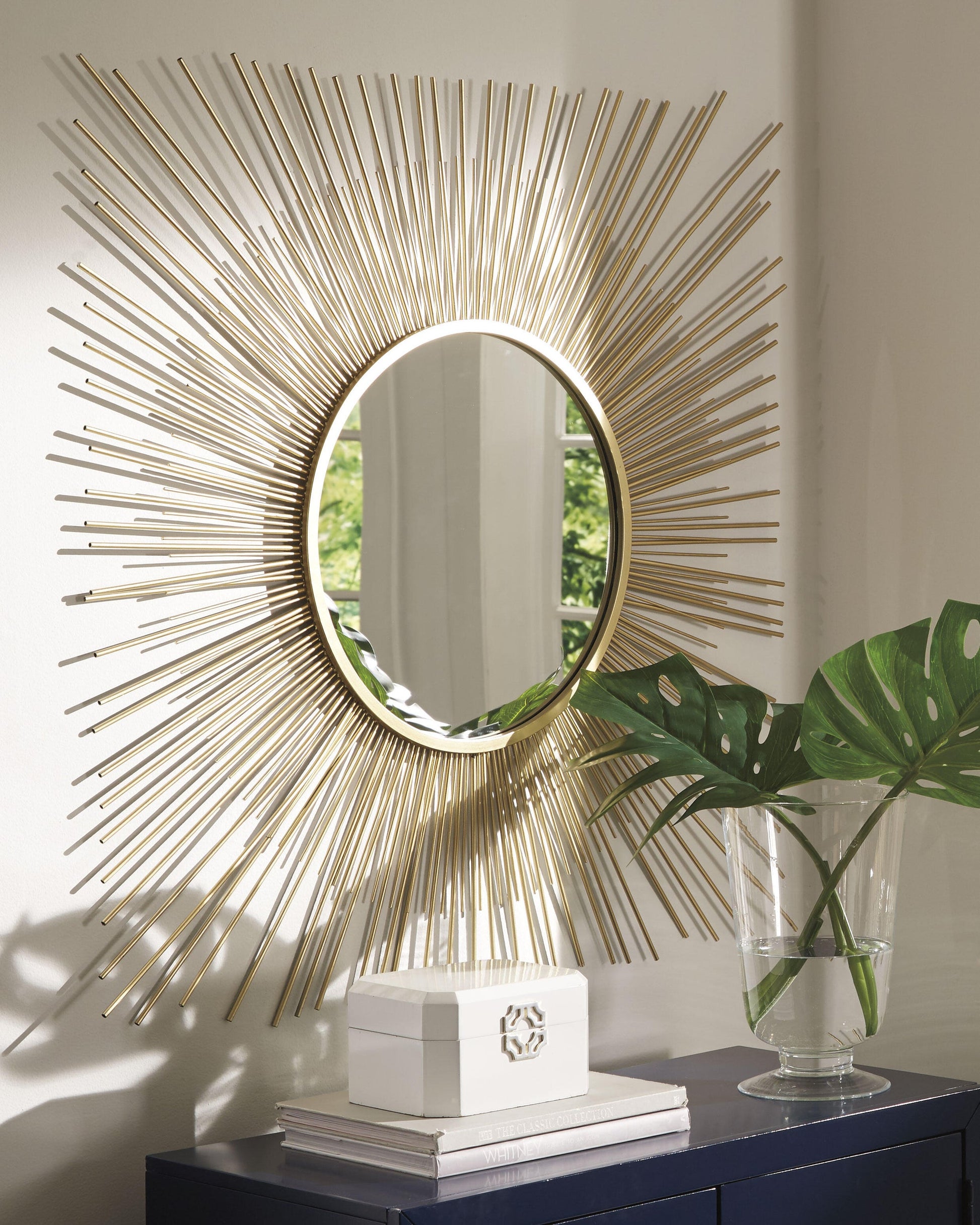 The Elspeth Accent Mirror, featuring a goldtone metal sunburst design, hangs above a dark cabinet beside a clear vase with green monstera leaves and a white decorative box.