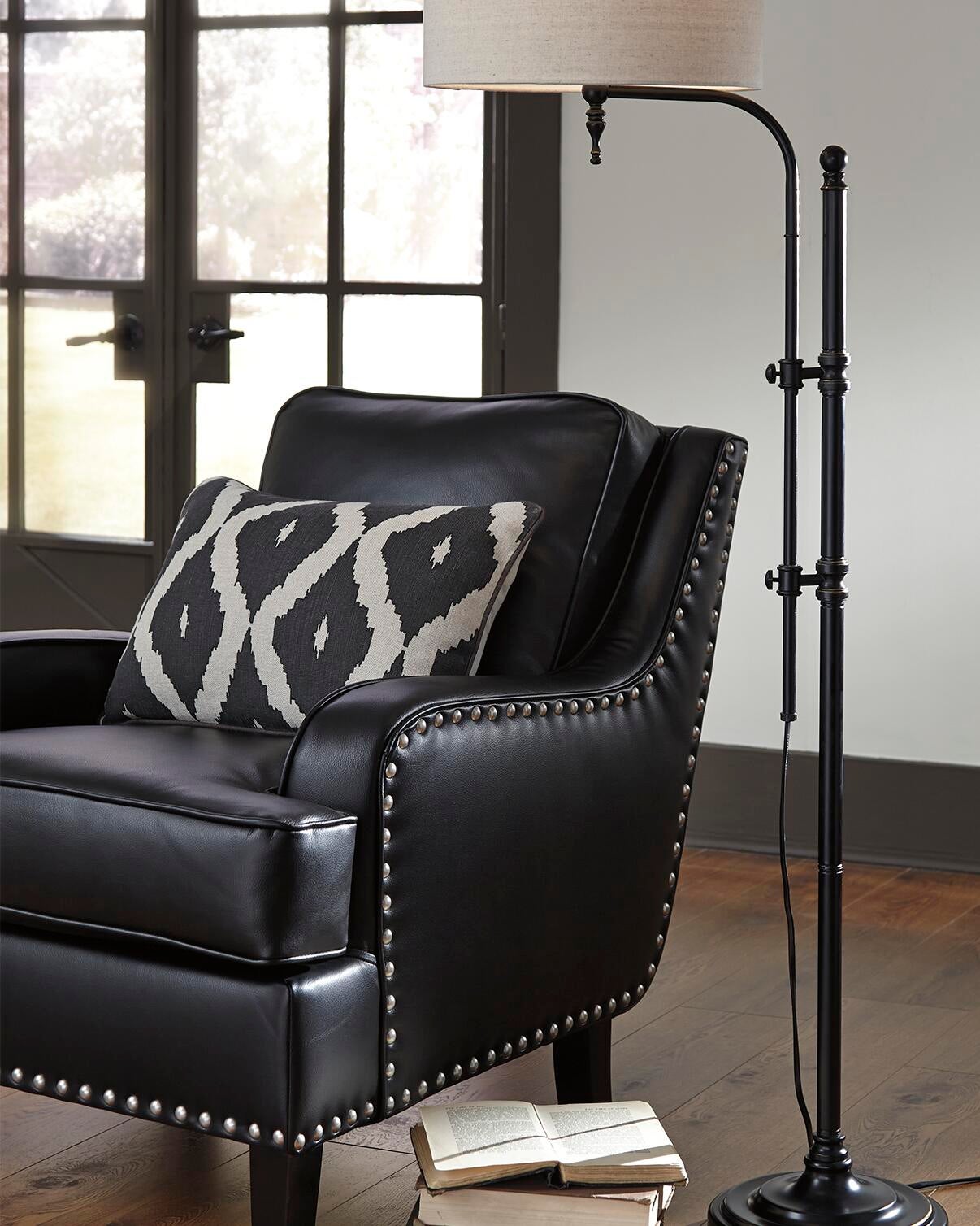 A black leather armchair with metal studs and a patterned cushion sits beside the Anemoon Metal Floor Lamp, which has an adjustable arm and metal base; books are stacked on the wooden floor near large windows in the background.