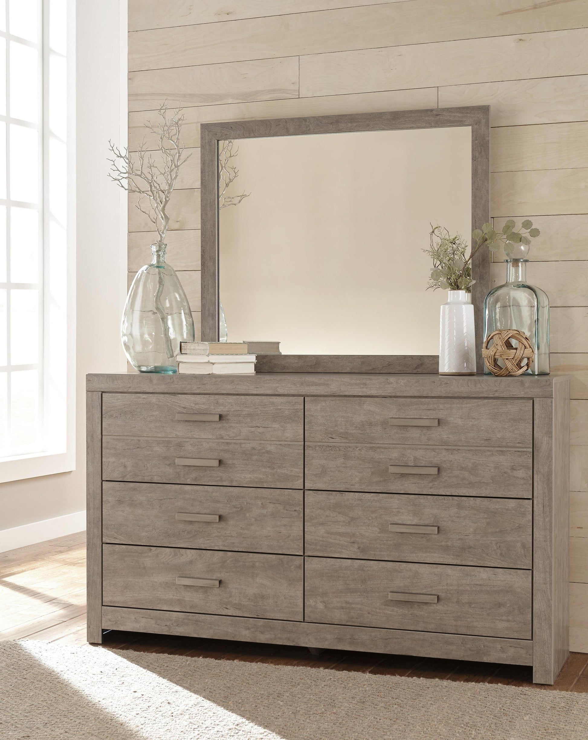The Culverbach Dresser and Mirror, finished in weathered driftwood, offers six spacious drawers and stands against a light wood-paneled wall, topped with glass vases, books, and a small plant.