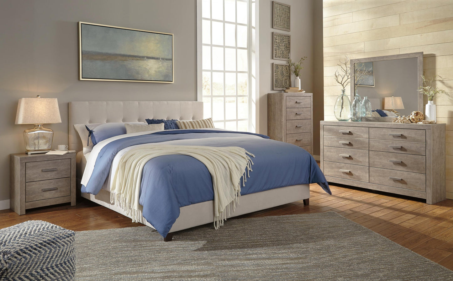 Modern bedroom with a queen bed featuring blue bedding, a beige headboard, two nightstands, and the Culverbach Dresser and Mirror in a weathered driftwood finish. Decorative art completes the look.