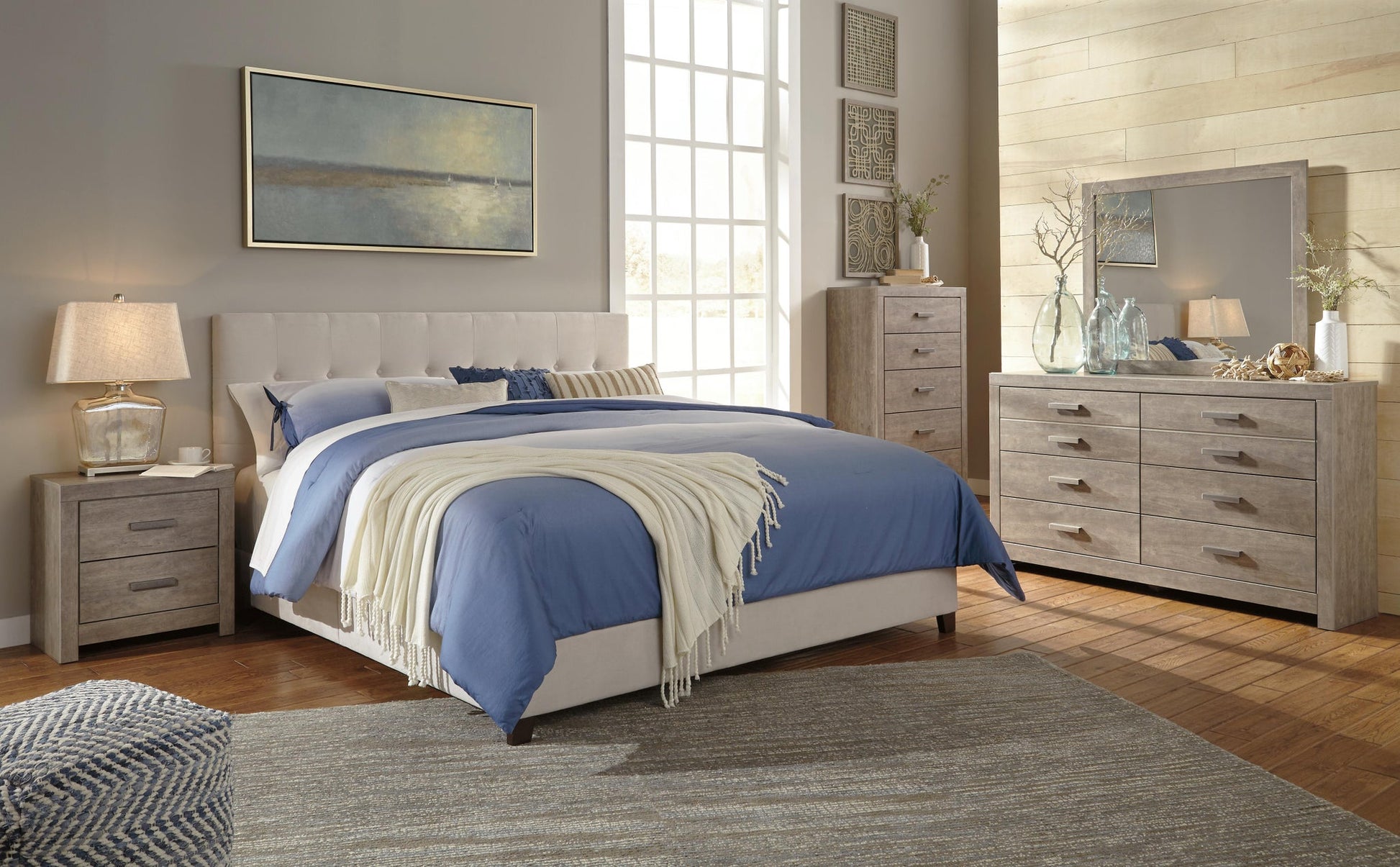 Modern bedroom with a queen bed featuring blue bedding, a beige headboard, two nightstands, and the Culverbach Dresser and Mirror in a weathered driftwood finish. Decorative art completes the look.