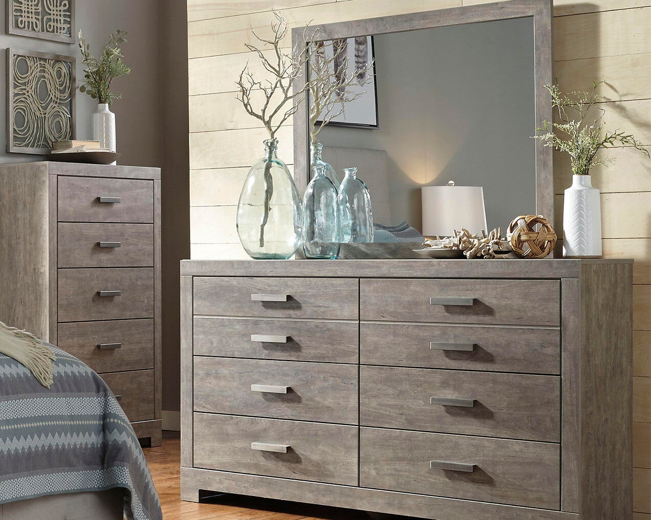 A bedroom showcases the Culverbach Dresser and Mirror in a weathered driftwood finish, six roomy drawers, a matching tall chest, glass vases, a lamp, plants, neutral colors, and geometric decor for a stylish look.