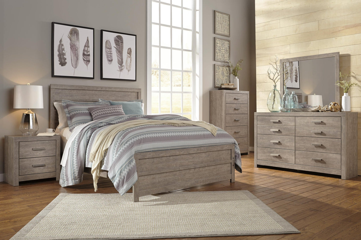 Modern bedroom with light wood furniture, patterned bedding, large window, nightstand, and feather wall art. The Culverbach Dresser and Mirror offer a weathered driftwood finish and six spacious drawers for ample storage.