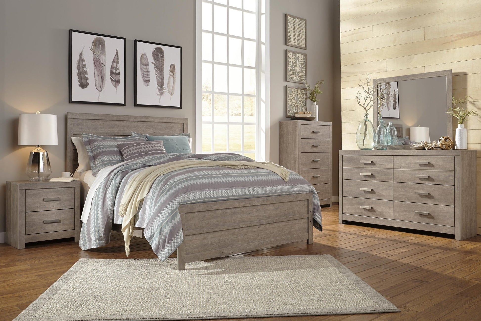 Modern bedroom with light wood furniture, patterned bedding, large window, nightstand, and feather wall art. The Culverbach Dresser and Mirror offer a weathered driftwood finish and six spacious drawers for ample storage.
