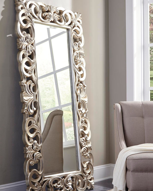 The Lucia Floor Mirror, featuring an intricate scroll design and antique silver finish, leans against a light wall near a window and sits beside a beige upholstered chair draped with a blanket.