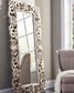 The Lucia Floor Mirror, featuring an intricate scroll design and antique silver finish, leans against a light wall near a window and sits beside a beige upholstered chair draped with a blanket.