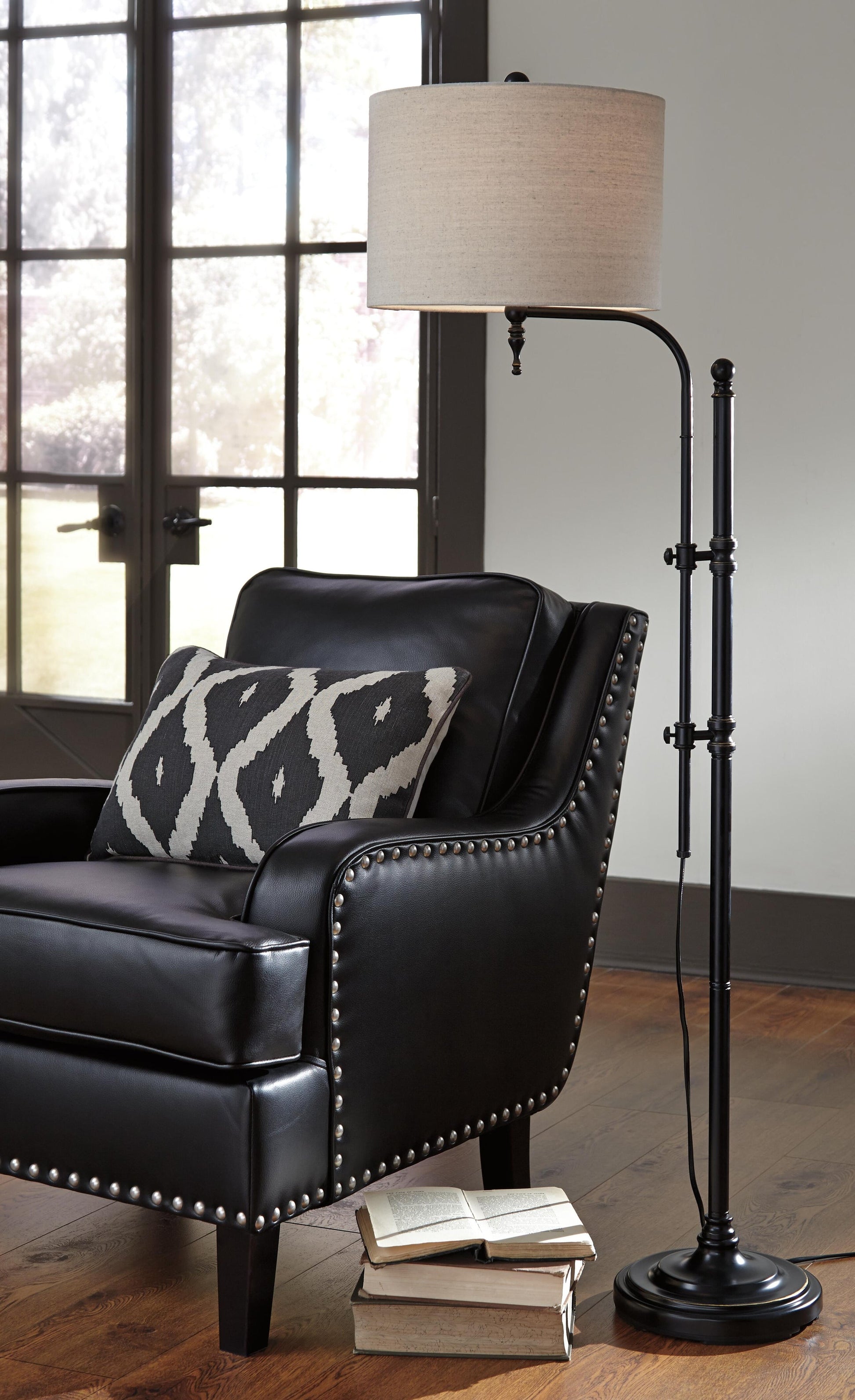 A black leather armchair with silver studs, a patterned cushion, and two open books on the wooden floor sits near tall windows, accompanied by the Anemoon Metal Floor Lamp with a metal base and adjustable arm.