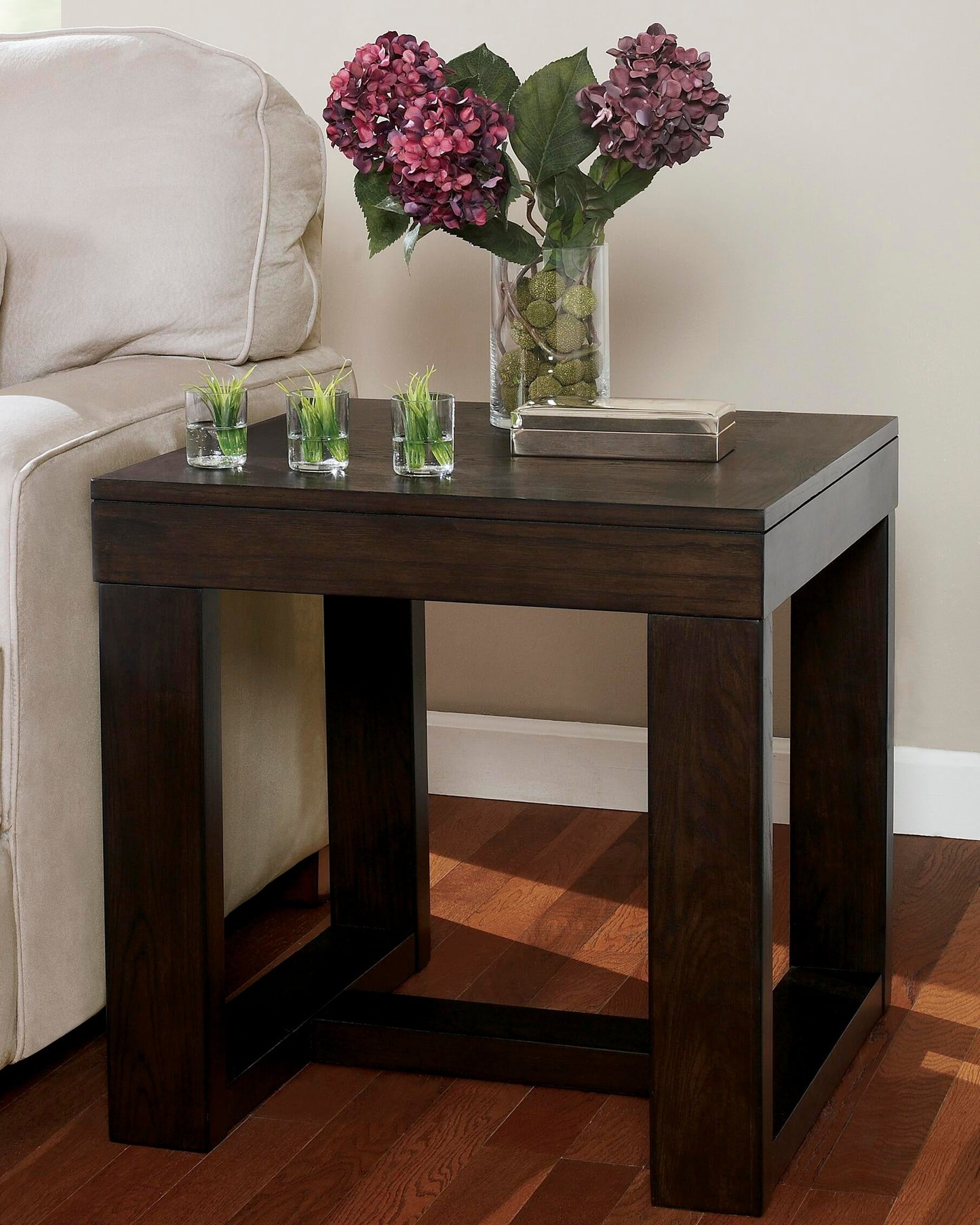 The Watson Square End Table sits by a beige sofa, topped with a glass vase of purple hydrangeas, a closed book, and three small square planters.