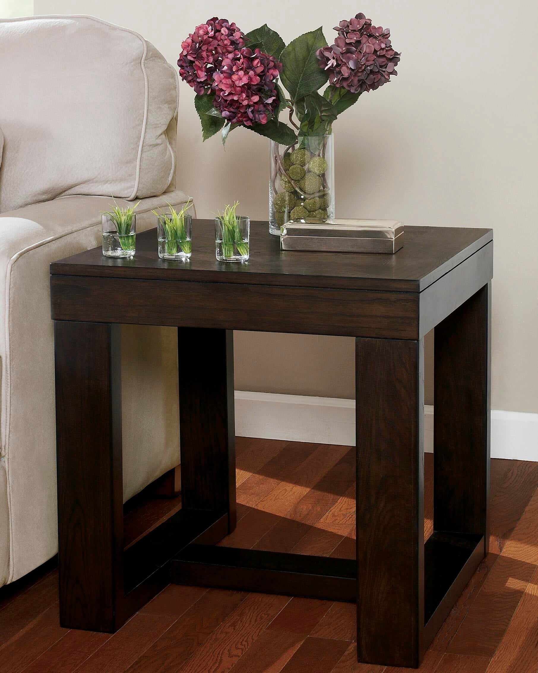 The Watson Square End Table sits by a beige sofa, topped with a glass vase of purple hydrangeas, a closed book, and three small square planters.