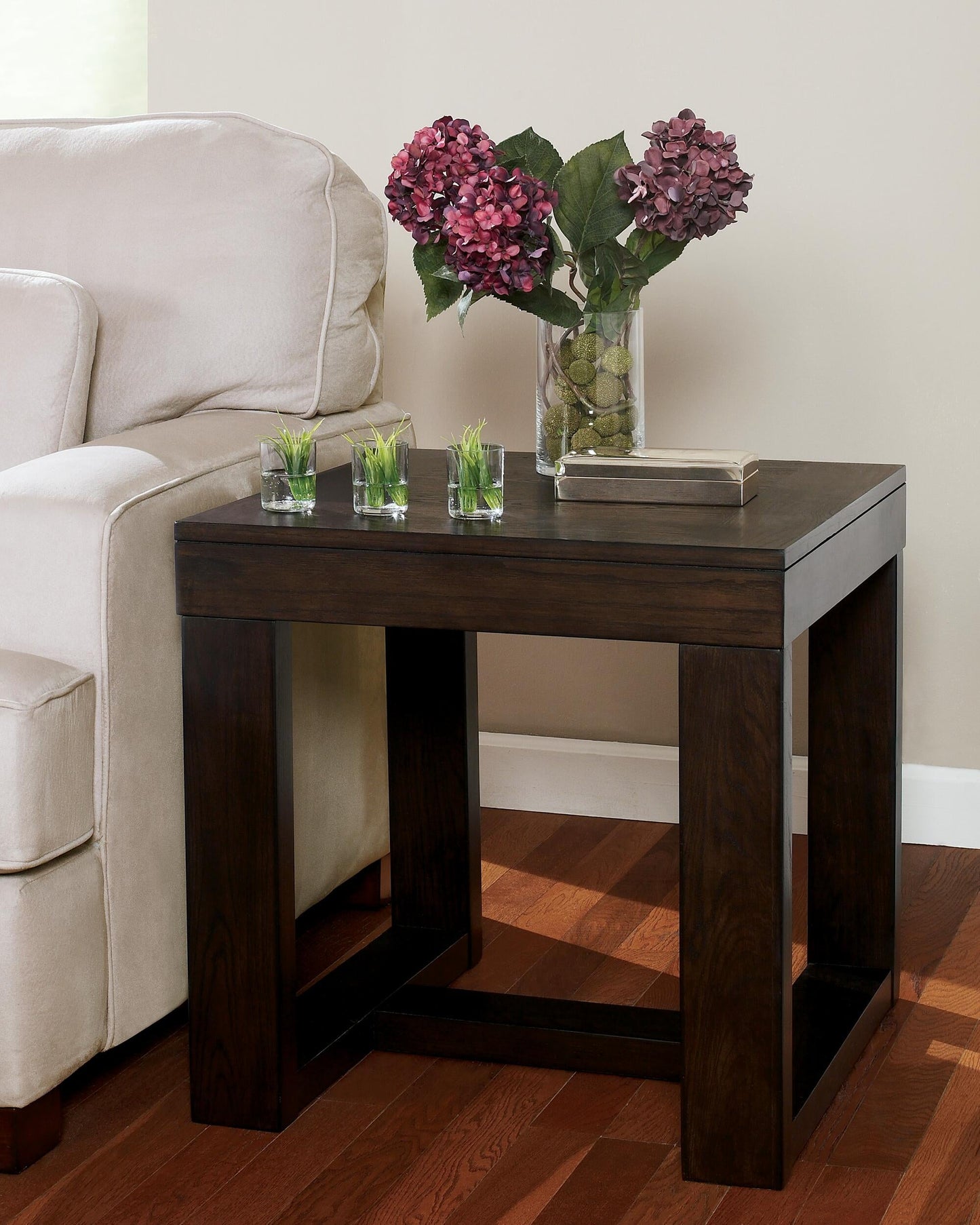 A Watson Square End Table beside a beige sofa holds three small glass planters with grass, a rectangular tray, and a vase of purple hydrangeas.